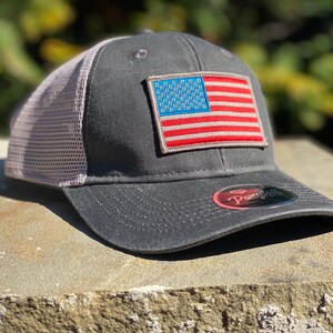 Puede incluir: Gorra de béisbol gris con parte trasera de malla y visera curva. La parte delantera de la gorra presenta un parche de la bandera estadounidense. La gorra tiene un ajuste estructurado y una visera curva. La parte trasera de malla es de color beige claro.