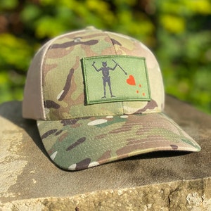 Puede incluir: Una gorra de camionero de camuflaje con un parche que muestra una calavera de pirata y huesos cruzados con un corazón rojo. La gorra tiene una parte trasera de malla y una visera marrón.