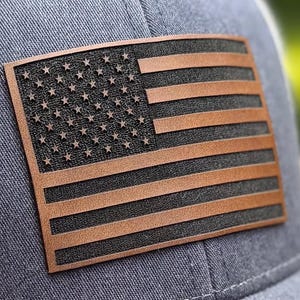 May include: A grey baseball cap featuring a rectangular patch on the front. The patch displays a stylized American flag design with raised brown leather-like stripes and stars set against a textured black background, stitched onto the fabric.