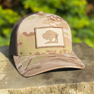 May include: A brown and tan camouflage trucker hat with a brown patch featuring a silhouette of a buffalo. The patch is sewn onto the front of the hat.