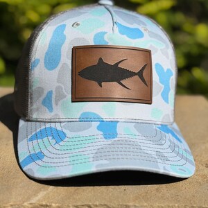 May include: A baseball cap featuring a light blue, white, and grey camouflage pattern. The front panel displays a rectangular brown leather patch with a black silhouette of a tuna fish centered on the patch. The cap has a curved brim and stitching.