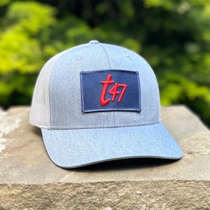May include: A light blue trucker hat with a white mesh back and a curved brim. The hat features a navy blue patch with the red text "t47". The hat is sitting on a stone surface.