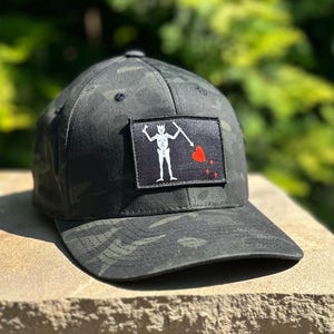 May include: A camouflage baseball cap with a black patch featuring a white skeleton holding a flag and a red heart. The cap has a curved brim and a structured fit. The hat is made of a durable fabric.
