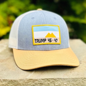 May include: A grey and white trucker hat with a yellow patch that says "TRUMP 45 * 47" and a blue and white flag with yellow mountains. The hat has a tan brim.