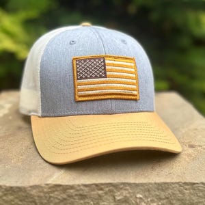 May include: A grey and white trucker hat with a brown and yellow American flag patch on the front. The hat has a tan coloured brim.