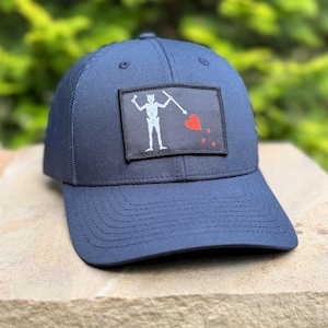May include: A navy blue baseball cap featuring a rectangular patch on the front. The patch displays a white skeleton figure holding a spear pointed toward a red heart, with three small red dots positioned below the heart on a black background.