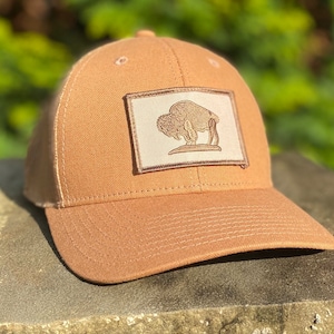 Könnte beinhalten: Eine beige Basecap mit einem braunen gestickten Aufnäher eines Büffels auf weißem Hintergrund. Der Aufnäher befindet sich auf der Vorderseite der Kappe.