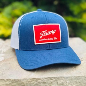 May include: A blue and white trucker hat with a red patch that says "Trump Smoother the 2nd Time" in white lettering.