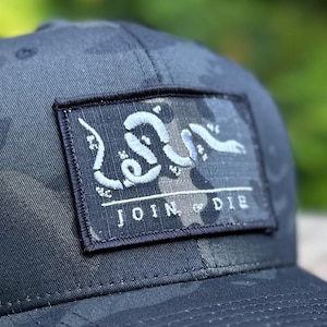 Puede incluir: Una gorra de camionero de camuflaje negro y gris con un parche negro que dice "JOIN OR DIE" y un diseño de serpiente bordado en blanco.