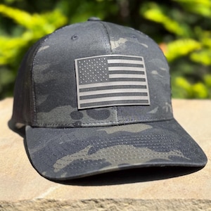 May include: A black and dark gray camouflage baseball cap featuring a rectangular patch on the front panel. The patch displays a monochromatic, embossed American flag design with stars and stripes in a muted, dark gray tone against a black background.