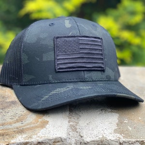 Puede incluir: Una gorra de camionero de camuflaje negro y verde con un parche de bandera estadounidense bordado en negro en la parte delantera.