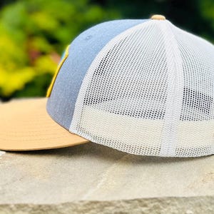 Puede incluir: Una gorra de camionero azul y tostada con una malla blanca en la parte posterior. La gorra tiene un dise&ntilde;o bordado amarillo en la parte delantera.