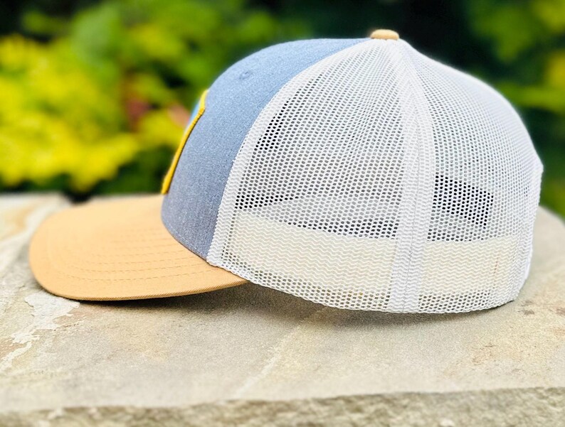 Puede incluir: Una gorra de camionero azul y tostada con una malla blanca en la parte posterior. La gorra tiene un dise&ntilde;o bordado amarillo en la parte delantera.