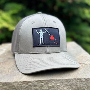 May include: A gray baseball cap with a black and white embroidered patch of a skeleton holding a spear and a red heart. The patch also has three red dots.