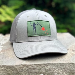 May include: A grey baseball cap with a mesh back and a curved peak. The front has a green patch with a black skeleton figure holding a spear and a red heart design. The hat is on a stone surface.