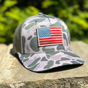 Puede incluir: Gorra de béisbol de camuflaje con parte trasera de malla y visera curva. La gorra presenta un parche de la bandera estadounidense en la parte delantera. El patrón de camuflaje incluye tonos de gris, verde y blanco.