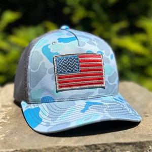 May include: A baseball cap featuring a blue camouflage pattern in shades of light blue, medium blue, and white. The front panel displays a rectangular embroidered patch of the United States flag with red, white, and blue stitching and a grey border.