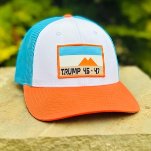 May include: A white and blue trucker hat with an orange brim. The hat has a patch on the front that says "TRUMP 45 * 47" and features a stylized mountain range.