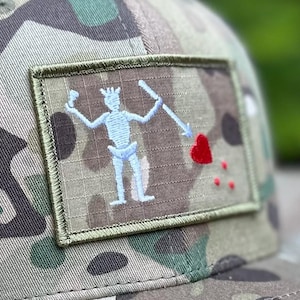 May include: A camouflage baseball cap with a green, brown and white pattern. The cap features a patch with a white skeleton holding a spear and a red heart. The patch has a green border.