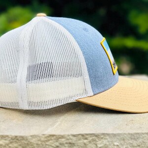 Puede incluir: Una gorra de camionero azul y blanca con una visera de color canela. La gorra tiene una malla blanca en la parte posterior y un parche en la parte delantera con un gr&aacute;fico de una cordillera.