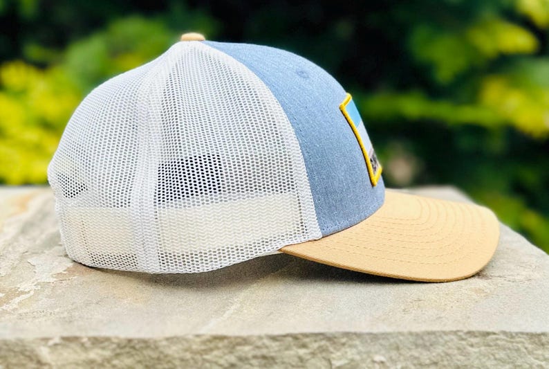 Puede incluir: Una gorra de camionero azul y blanca con una visera de color canela. La gorra tiene una malla blanca en la parte posterior y un parche en la parte delantera con un gr&aacute;fico de una cordillera.