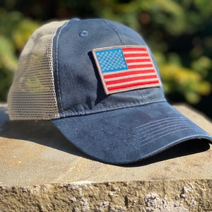 Può includere: Un cappello da baseball blu e beige con una rete sul retro e una toppa della bandiera americana rossa, bianca e blu sul davanti.