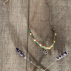 May include: A handcrafted necklace featuring a brown cord, green and yellow beads, and a silver four-leaf clover charm. Two lavender sprigs are crossed beneath the necklace, set against a wooden backdrop.
