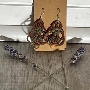 May include: A pair of handmade macrame earrings with a leaf design. The earrings are brown with small green and yellow beads. They hang from silver-colored hooks and are displayed on a brown tag. Lavender sprigs are placed on either side of the earrings.