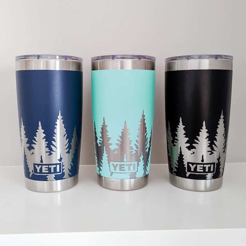 Pode incluir: Tr&ecirc;s copos YETI com um design de silhueta de floresta. Os copos s&atilde;o azul marinho, azul-turquesa e preto.