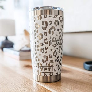 Peut inclure: Un gobelet YETI en acier inoxydable avec un extérieur blanc orné d'un motif léopard métallique. Le récipient cylindrique possède un couvercle transparent et une base argentée, avec la marque YETI gravée dans un cadre rectangulaire en bas.