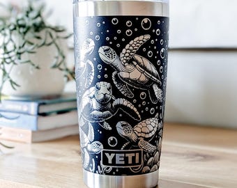 May include: A black stainless steel tumbler with a clear lid, featuring a white engraved pattern of sea turtles swimming among bubbles. The YETI brand logo is printed in white at the bottom center of the cylindrical vessel, which has a silver base.