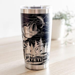 Laser-engraved stainless steel Yeti tumbler featuring a camping and fishing design, including an outdoor pine forest landscape and a person fishing from a boat on a lake.