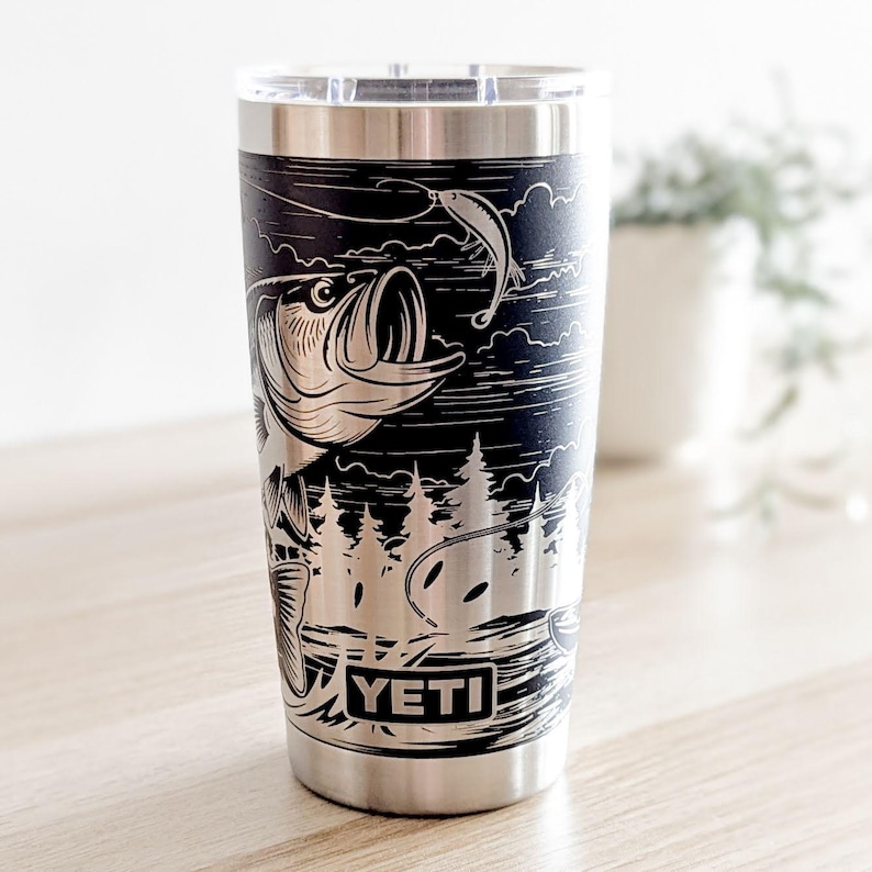 Laser-engraved stainless steel Yeti tumbler featuring a camping and fishing design, including an outdoor pine forest landscape and a person fishing from a boat on a lake.