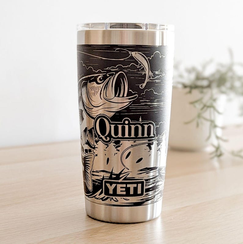 Laser-engraved stainless steel Yeti tumbler featuring a camping and fishing design, including an outdoor pine forest landscape and a person fishing from a boat on a lake.