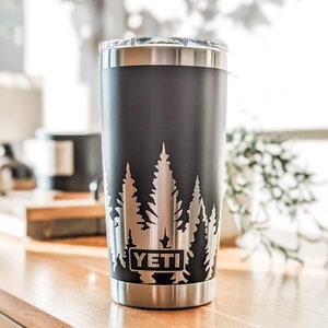 Pode incluir: Um copo YETI preto com tampa prateada e base prateada. O copo tem um design de &aacute;rvores gravadas no lado.