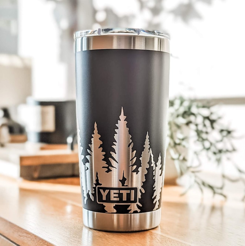 Pode incluir: Um copo YETI preto com tampa prateada e base prateada. O copo tem um design de &aacute;rvores gravadas no lado.