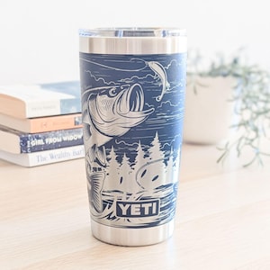 May include: A navy blue stainless steel tumbler featuring a laser-etched graphic of a large fish jumping out of water toward a fishing lure, with a forest background and the YETI brand name printed in a rectangular frame at the bottom base.