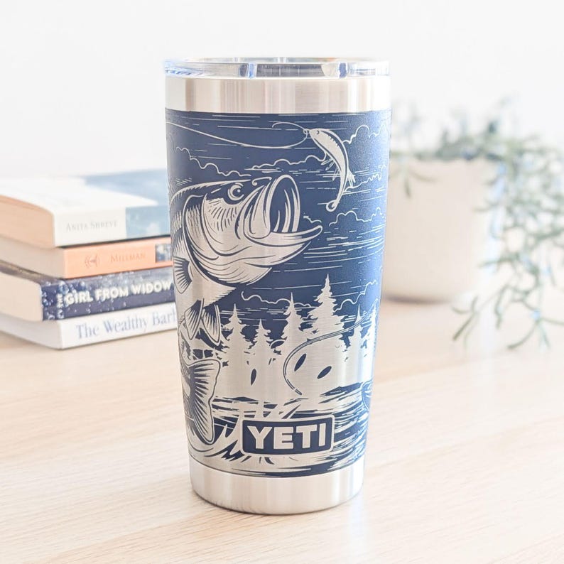 May include: A navy blue stainless steel tumbler featuring a laser-etched graphic of a large fish jumping out of water toward a fishing lure, with a forest background and the YETI brand name printed in a rectangular frame at the bottom base.