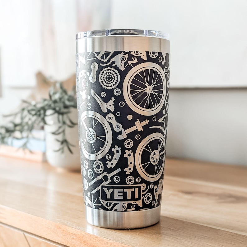 May include: A silver Yeti tumbler with a black background and a silver design of bicycle parts, including wheels, gears, and chains. The word "YETI" is printed in silver on the tumbler.