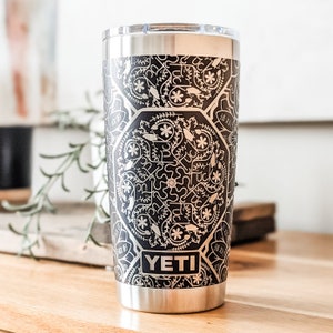 May include: A black and white Yeti tumbler with a geometric pattern. The tumbler has a stainless steel exterior and a black lid. The word "YETI" is printed in white on the front of the tumbler.