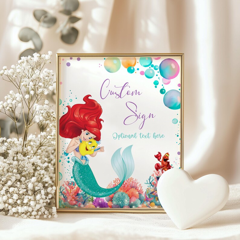 May include: A framed custom sign featuring a mermaid with red hair and a teal tail, holding a yellow fish. The sign includes the text "Custom Sign" and "Optional text here" with colorful bubbles in the background. A white heart-shaped object is in the foreground.