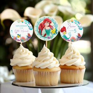 May include: Three cupcakes with white frosting, each topped with a round paper decoration. The decorations feature images of Ariel from The Little Mermaid, along with text that reads "Happy Birthday to our Little Mermaid" and "Siena is Turning 5".
