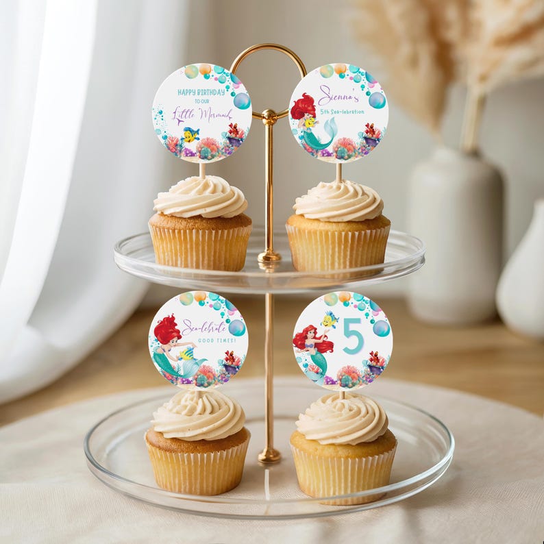 May include: A two-tiered dessert stand holds four cupcakes, each topped with a swirl of frosting. Each cupcake has a round topper with a mermaid theme, featuring text like "Happy Birthday" and "Little Mermaid". The toppers have a mermaid and sea life illustrations.