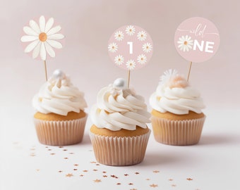 May include: Three white cupcakes with pink and white daisy themed cupcake toppers. The toppers say "wild one" and "1" surrounded by daisies. The cupcakes are on a white surface with gold glitter stars.