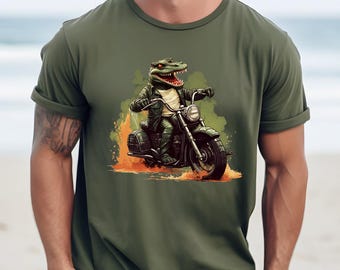 May include: Olive green t-shirt featuring a cartoon alligator riding a motorcycle. The alligator is wearing a leather jacket and helmet. The motorcycle is black with flames around the tires. The background is a beach scene.