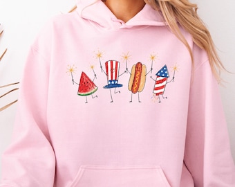 May include: A light pink hooded sweatshirt featuring a graphic print of a smiling watermelon slice, a top hat with stars and stripes, a hot dog, and a firework rocket, all holding sparklers to celebrate the Fourth of July holiday.