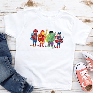 May include: White t-shirt featuring a cartoon illustration of superheroes, including Spider-Man, Iron Man, Hulk, Captain America, and others. The shirt is paired with blue jeans and coral-colored sneakers. A small potted plant is also visible.