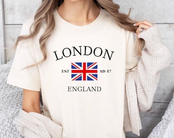 May include: Cream-colored t-shirt with the word "LONDON" in black, above a Union Jack flag graphic. Below the flag are the words "EST AD 47 ENGLAND" in black. The shirt is short-sleeved.