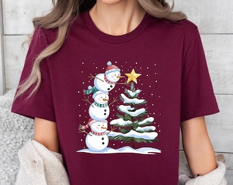May include: Burgundy t-shirt featuring a whimsical Christmas design. The graphic depicts a snowman stack next to a snow-covered Christmas tree, topped with a golden star. The snowmen wear hats and scarves. A festive holiday tee.
