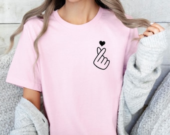 May include: A pink t-shirt with a black hand gesture and a small heart on the front. The hand gesture is a popular Korean symbol for love.